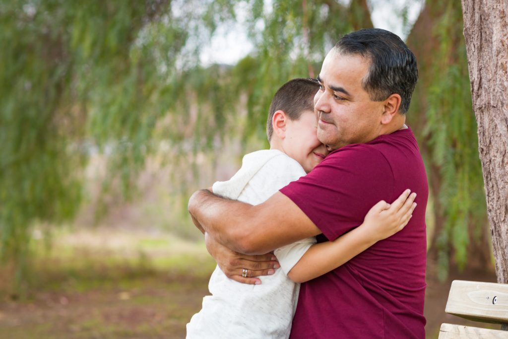 Foto mostra um homem e um menino se abraçando, felizes, em um parque.