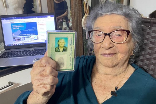 Foto da eleitora Maria Guiomar Tavares Ferreira, de 106 anos. Ela usa óculos e tem cabelos brancos, usa blusa azul e segura o documento de identidade.