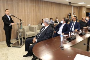 Foto mostra momento do evento, com um homem em pé falando em um microfone e participantes sentados em volta de uma mesa olhando para ele.