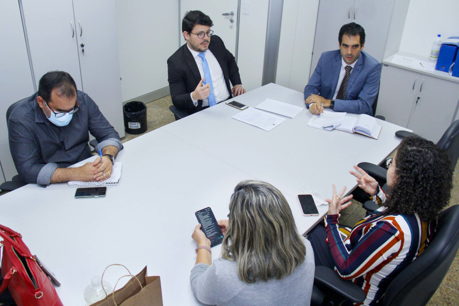 Foto mostra cinco pessoas sentadas em volta de uma mesa de reunião em um escritório. São três homens e duas mulheres conversado.