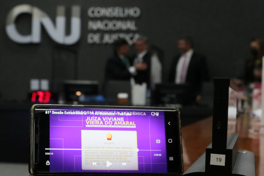 Foto mostra em destaque à frente um celular na posição horizontal mostrando trecho da apresentação dos ganhadores do Prêmio CNJ Juíza Viviane Vieira do Amaral. Ao fundo, desfocado, percebe-se o Plenário do CNJ com pessoas se cumprimentando.