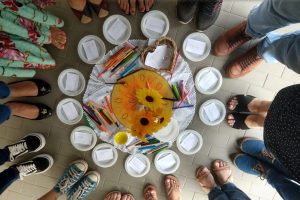 Foto mostra pés de pessoas, em círculo, em volta de papeis, canetas e lápis para realização de atividade restaurativa.