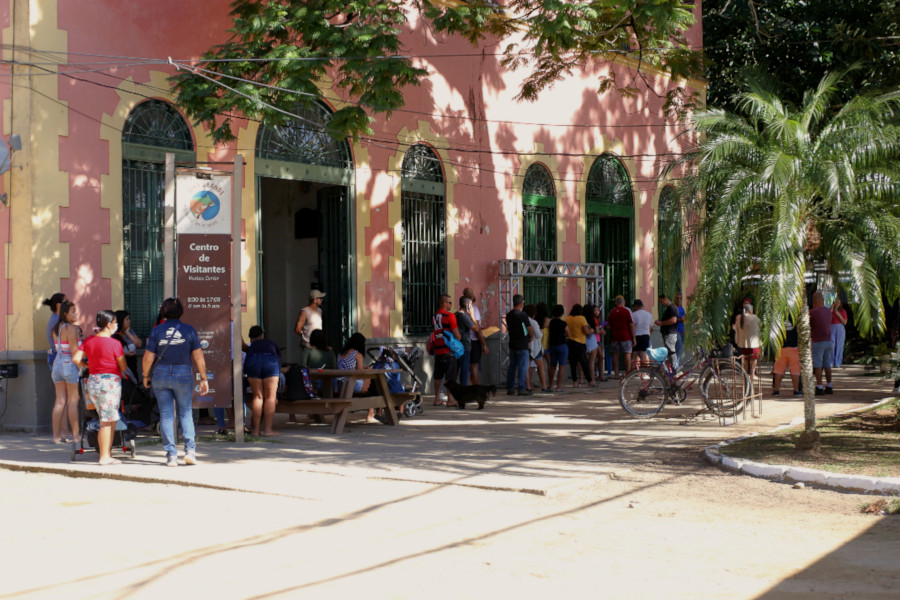 Foto mostra a área externa da Casa de Cultura, com diversas pessoas aguardando em fila para serem atendidas.