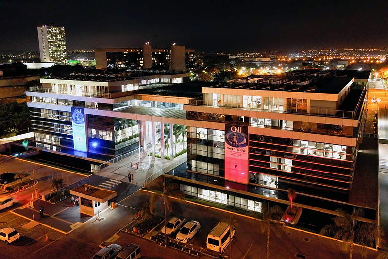 Foto noturna da sede do CNJ, com a fachada de um prédio iluminada de azul e a do outro prédio de rosa.