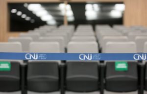 Fotografia em plano detalhe: Fita de cetim em tom azul claro e nela está a logo do CNJ - Conselho Nacional de Justiça. Ao fundo embaçado cadeiras do plenário do CNJ.
