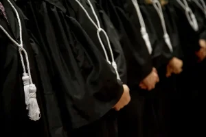 Imagem em plano detalhe, magistrados enfileirados usando Toga ( traje usado nos tribunais) de tom escuro e cordão de tom prata.