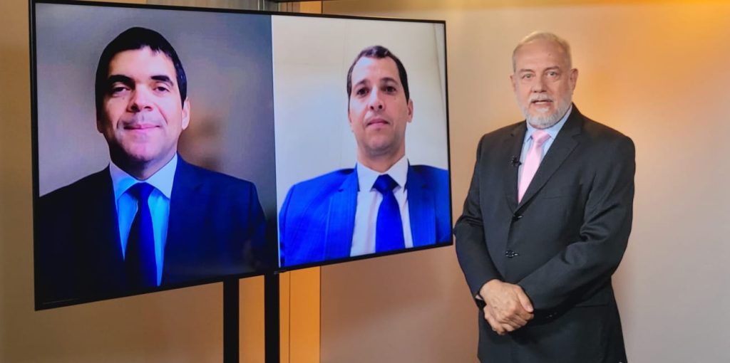 Foto do estúdio do programa com o apresentador posando ao lado do telão onde se vê as pessoas entrevistadas por videoconferência.