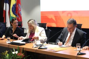 Fotografia em questão em destaque a procuradora-geral do Estado da Bahia, Bárbara Camardelli; a presidente do TJBA, desembargadora Cynthia Maria Pina Resende; e do presidente do Conselho Nacional de Justiça (CNJ) e do Supremo Tribunal Federal (STF), ministro Luís Roberto Barroso enquanto assinam um documento. No canto superior esquerdo, há três bandeiras. No canto inferior esquerdo, há algumas flores de cores variadas como branca e vermelha.