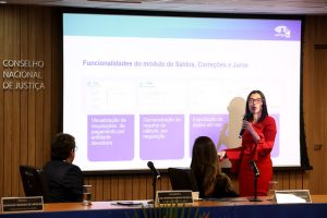 Fotografia da Juíza auxiliar da Presidência do CNJ, Wanessa Mendes de Araújo falando em um microfone, ela está na frente de uma tela de projeção. Ela veste um vestido vermelho, têm cabelos pretos curtos e usa óculos de armação vermelha. No canto esquerdo da foto, na parede, está inscrição “Conselho Nacional de Justiça”. Em frente a ela, um homem e uma mulher a observam atentamente. Eles estão sentados atrás de uma mesa.