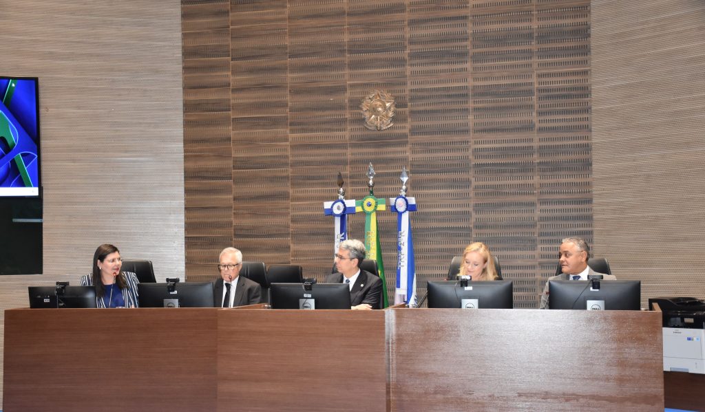 Fotografia do Tribunal Regional Federal da 2ª Região no Rio de Janeiro (TRF2), onde estão presentes o presidente do TRF2, desembargador federal Guilherme Calmon (que usa um terno cinza escuro, uma gravata azul, e óculos), a conselheira do CNJ, Daiane Nogueira de Lira (de blusa azul, blazer de listras azuis e brancas e óculos), a coordenadora do Comitê Estadual do Fórum Nacional de Saúde do CNJ no Rio de Janeiro, desembargadora federal Carmen Silva Lima de Arruda (de blusa branca e blazer preto com detalhes pequenos de flores azuis e brancas), o presidente do Tribunal Regional do Trabalho da 1ª Região (TRT1), desembargador Cesar Marques Carvalho (de terno cinza escuro, gravata preta e óculos) e o juiz federal Roberto Schuman, membro do Comitê Estadual de Saúde (de terno cinza e gravata azul). Eles estão sentados atrás de uma mesa, que possui computadores em cima. Acima deles, está o brasão do Brasil. 