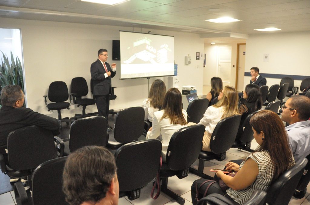 Fotografia de pessoas sentadas em uma sala participando do projeto “Café com Cejusc”. Ao centro, um homem de terno fala se direcionando as pessoas que estão em sua frente. No canto direito, ao lado do homem, uma televisão transmite uma imagem