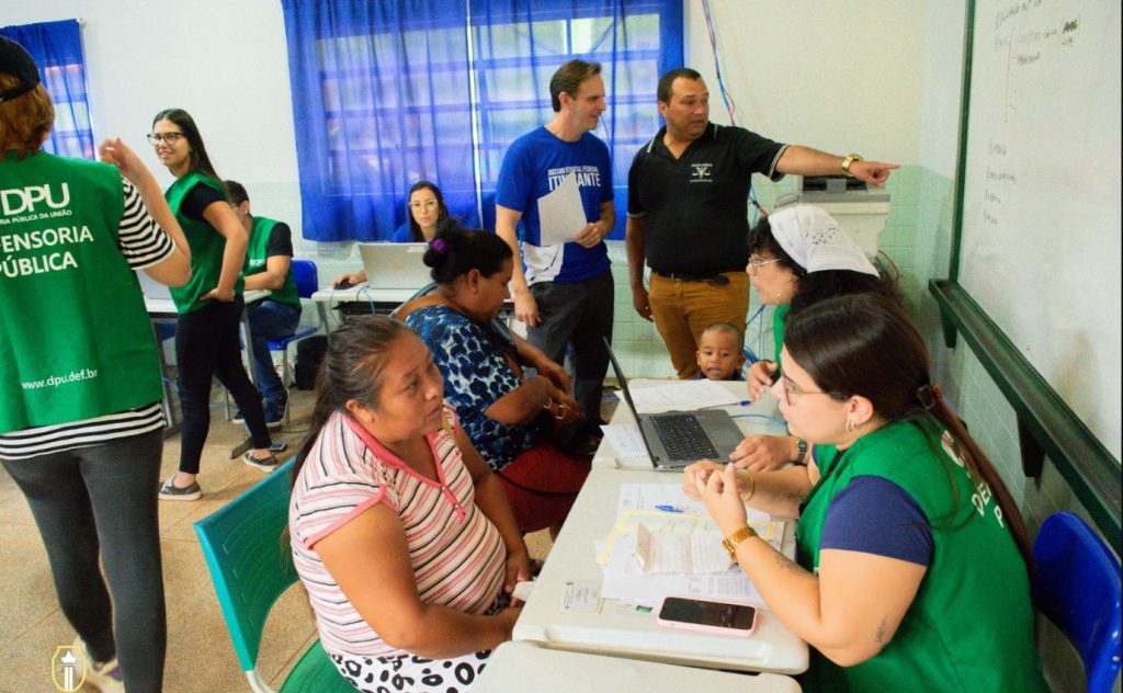 Fotografia em que há várias pessoas em uma sala que parece ser de uma escola. Algumas estão de colete verde com inscrição da Defensoria Pública conversando com outras pessoas que parecem se informar 