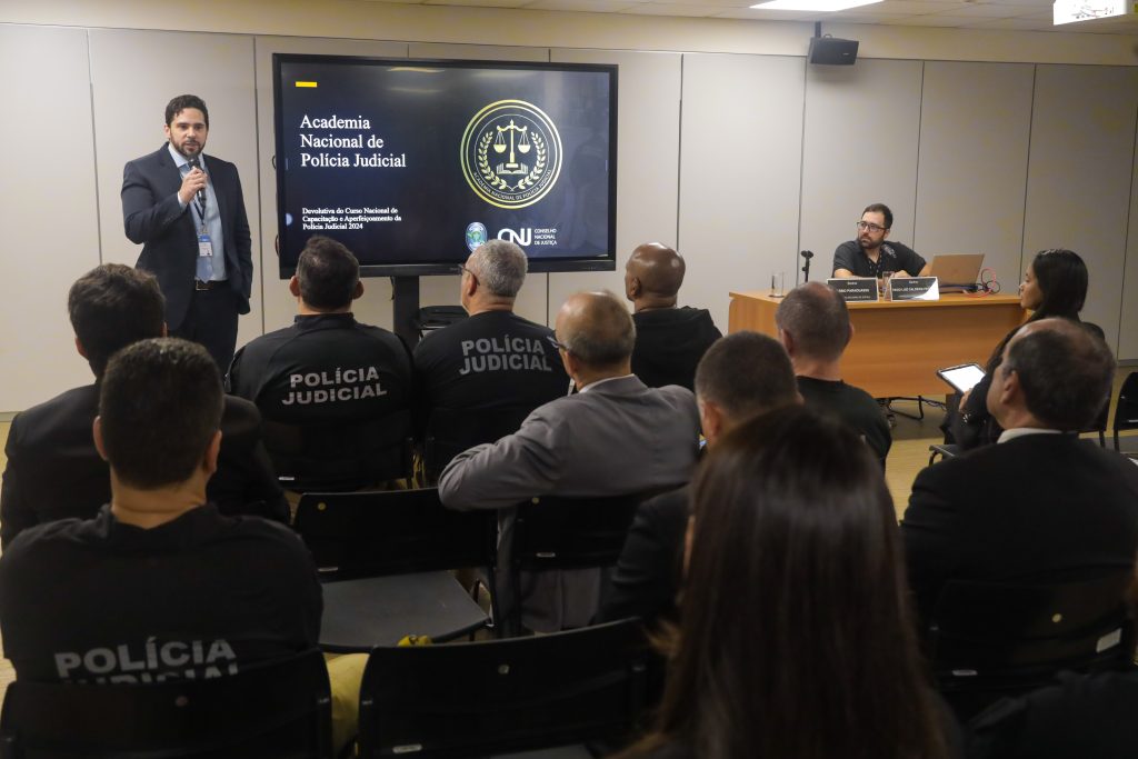 Fotografia de diversas pessoas em uma sala de reunião. Pessoas assistem apresentação de um homem de terno está de pé e fala em um microfone. Ao lado direito do homem, há uma televisão que apresenta texto: Academia Nacional de Polícia Judicial, do lado direito, o símbolo da academia e na parte de baixo, a sigla do Conselho Nacional de Justiça (CNJ).  