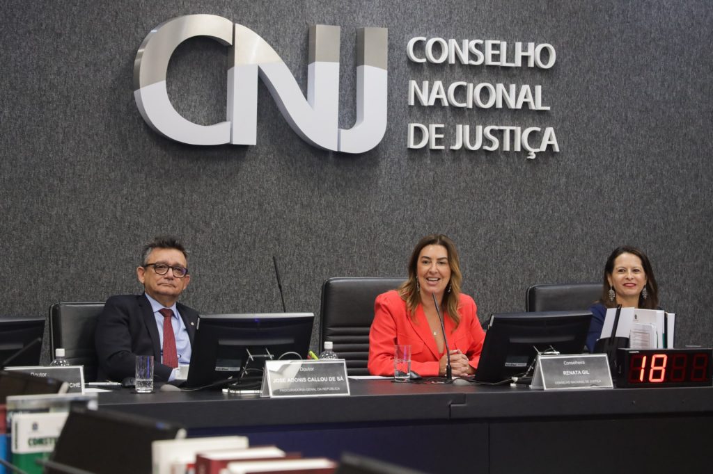 Fotografia do Dr. José Adonis Callou de Sá da Procuradoria Geral da República, da Conselheira Renata Gil, do CNJ e da Juíza Auxiliar da presidência do CNJ Luciana Lopes. Todos estão sentados. A conselheira Renata Gil fala em um microfone. O Dr. José de Sá usa um terno, com uma gravata vermelha. A conselheira Renata Gil usa um blazer laranja. A juíza auxiliar Luciana Lopes usa um blazer azul escuro e brincos pratas. 