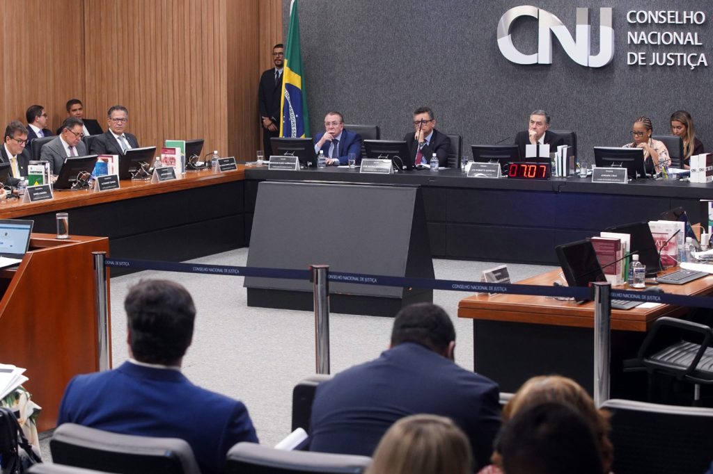 Fotografia do Plenário do Conselho Nacional de Justiça (CNJ). No canto superior direito, a sigla do Conselho Nacional de Justiça (CNJ). Do lado esquerdo, a bandeira o Brasil em um mastro. No centro, há uma mesa formada por doutores, além do presidente do Conselho Nacional de Justiça (CNJ) e do Supremo Tribunal Federal (STF), ministro Luís Roberto Barroso. Na parte inferior da imagem pessoas sentadas assistem à sessão ordinária.  