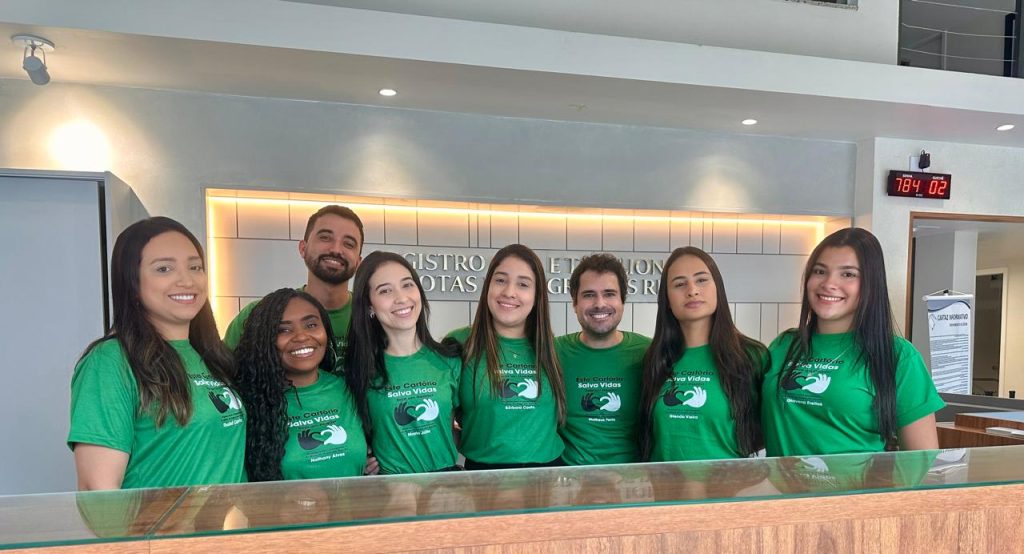 Fotografia da Equipe do Cartório de Registro Civil e Tabelionato de Notas de Angra dos Reis. A foto é composta por seis mulheres e dois homens, todos usam uma blusa verde com a logo da campanha Um Só Coração. Eles estão em pé atrás de um balcão. Ao fundo, em uma parede branca e rodeado por uma luz de led amarelada, está o letreiro com o nome do cartório.  