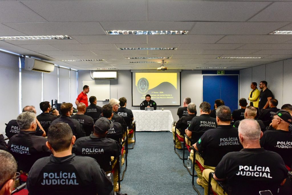 Fotografia de diversos policiais sentados em uma sala enquanto participam de um curso que está sendo administrado por um outro policial que está sentado atrás de uma mesa e em frente a uma tela de projeção. Todos usam uniformes da Polícia Judicial, que é composta por uma blusa preta com a inscrição ‘Polícia Judicial” na parte de trás da camisa, uma calça e botas de cor bege. 