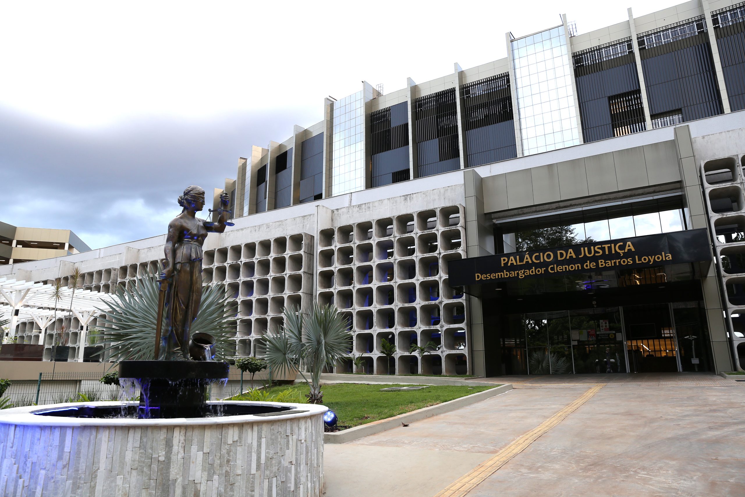 Fotografia da frente do Tribunal de Justiça de Goiás (TJGO). Na entrada, há a inscrição “Palácio da Justiça Desembargador Clenon de Barros Loyola”. Em frente à entrada, uma fonte de água, com uma estátua da Justiça, uma mulher vendada que segura uma balança e uma espadano, ao centro. Atrás da fonte, há algumas palmeiras de pequeno porte em um gramado. 