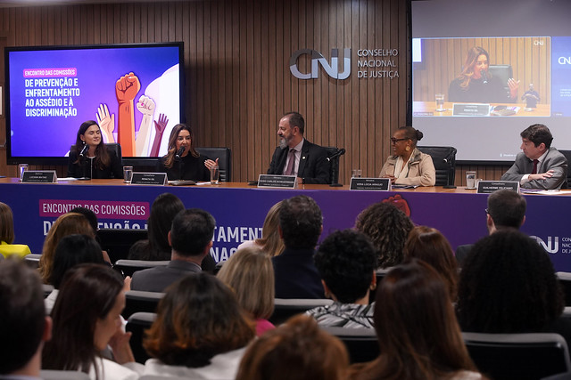 Fotografia de um evento onde estão (da esquerda para a direita): do Conselho Nacional de Justiça (CNJ), senhora Luciana Matias (mulher de cabelos lisos e castanhos, que veste uma blusa preta com uma listra branca, um blazer preto e brincos), conselheira Renata Gil (mulher de cabelos lisos e castanho claro, e veste uma blusa e blazer preto), do Tribunal Superior do Trabalho, Ministro Hugo Carlos Scheuermann (homem de cabelos e barba grisalhas, veste uma camisa social lilás, uma gravata vermelha e um terno preto), do Tribunal Superior Eleitoral, Ministra Vera Lúcia Araújo (mulher de cabelos presos em um penteado coque, usa óculos e brincos, e veste um blazer e um lenço bege) e também do CNJ, o conselheiro Guilherme Feliciano (homem de cabelos pretos, que veste camisa social branca, gravata vermelha, e terno cinza). Eles estão sentados atrás de uma mesa, e ao fundo há o logotipo do Conselho Nacional de Justiça (CNJ) na parede, juntamente com a inscrição “Conselho Nacional de Justiça”, uma televisão que transmite um card roxo com alguns tons rosa que apresenta a inscrição “Encontro das Comissões de Prevenção e Enfrentamento do Assédio e da Discriminação” no lado esquerdo. Do lado direito, desenhos de mãos de mulheres, alguns com punhos fechados e outras coma mão aberta, em vários tons de pele, e uma tela de projeção que transmite o evento. Algumas pessoas acompanham de maneira presencial, e estão sentadas em frente as pessoas que estão na mesa.