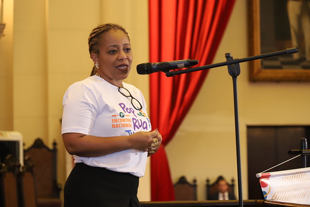 Fotografia da secretária-geral do CNJ, Adriana Cruz ((mulher negra de cabelos trançados loiros, usa brincos, e veste uma blusa branca com a estampa com uma inscrição nas cores roxa, azul e amarela, e apresenta o texto: “I Encontro Nacional do PopRuaJud” e uma saia preta. Ela tem óculos de armação preta pendurados na blusa.) Ela fala em um microfone. Ela está em uma sala espaçosa, com várias janelas e cortinas vermelhas. 