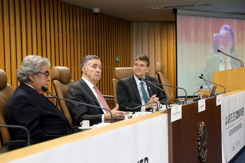 Fotografia do presidente do STF e do CNJ, ministro Luís Roberto Barroso (ao centro), do presidente do TST e do CSTJ, ministro Aloysio da Veiga (na esquerda) e do conselheiro do CNJ, Alexandre Teixeira. Eles estão em uma plenária no Tribunal de Justiça Social. O ministro Barroso (homem branco, veste uma camisa social branca, uma gravata vermelha com estampa de bolinhas e um terno preto) fala ao microfone, enquanto o ministro Veiga (homem branco de cabelos grisalhos, usa óculos de armação preta, e veste uma camisa social listrada, uma gravata azul com bolinhas brancas, e um terno azul marinho) e o conselheiro Teixeira (homem branco de cabelos claros, veste uma camisa social branca, gravata verde com bolinhas brancas e terno preto) o observam. Eles estão sentados atrás de uma mesa de madeira com o brasão do Brasil estampado na frente da mesa. No lado direito da fotografia, uma tela de projeção transmite a imagem do ministro Barroso. 