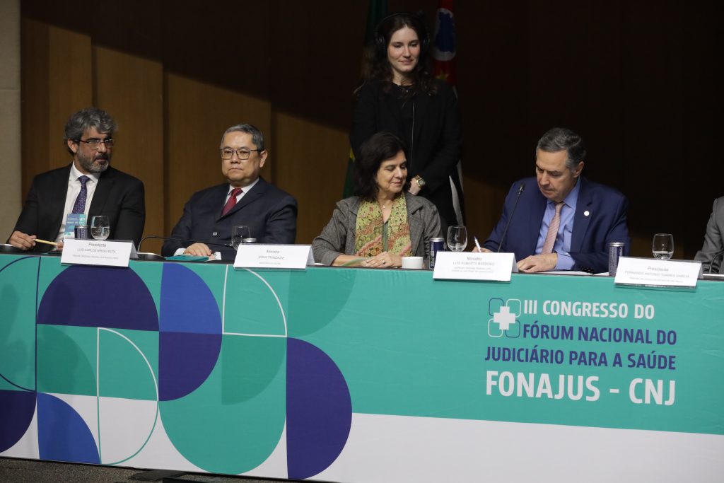 Presidente do Conselho Nacional de Justiça (CNJ) e do Supremo Tribunal de Justiça (STF), ministro Luís Roberto Barroso (de camisa social azul, gravata vermelha e terno azul) enquanto está sentado no centro de uma mesa verde com as inscrições: “III Congresso do Fórum Nacional do Judiciário para a Saúde”, “Fonajus - CNJ”. O logotipo do fórum aparece ao lado esquerdo das inscrições.  Ele assina um documento que está sobre a mesa. A mulher que está ao lado dele (de cabelos curtos e escuros, blusa verde, blazer cinza e lenço verde com estampa colorida) observa enquanto ele assina. Ao lado dela, há também um homem de cabelos e barba grisalha, óculos, veste camisa social branca, gravata roxa e terno preto; homem de cabelos grisalhos, usa óculos, e veste camisa social branca, gravata vermelha e terno preto. Atrás deles há uma mulher de cabelos longos, e que veste uma roupa preta, de pé. 