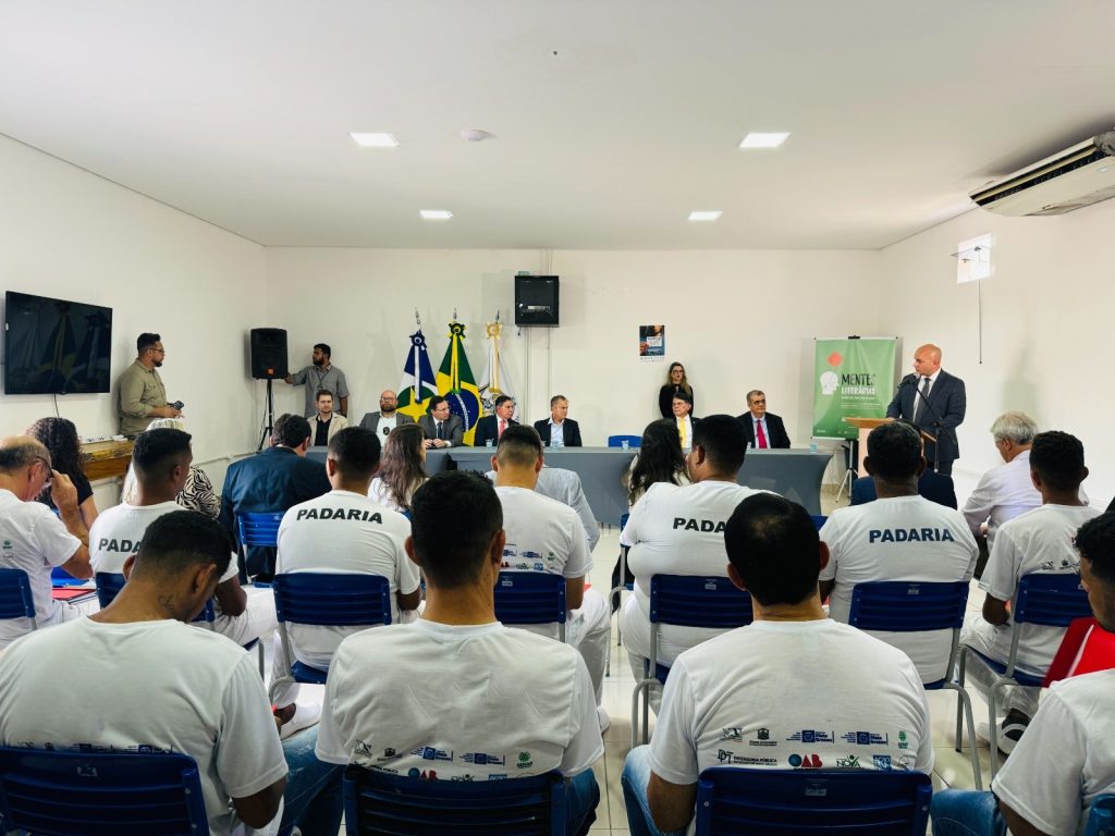 Fotografia onde aparecem onze pessoas, que vestem, camisas brancas e calças jeans azuis e estão sentadas e, cadeiras azuis, enquanto observam um homem que usa um terno azul escuro que está em um palanque, falar em um microfone. Ao lado do homem que está no palanque, há uma mesa com sete outros homens sentados. No canto esquerdo da foto, três bandeiras, sendo uma delas a do Brasil, estão em mastros. Há também um banner verde ao fundo, com a inscrição: “Mentes Literárias”. 