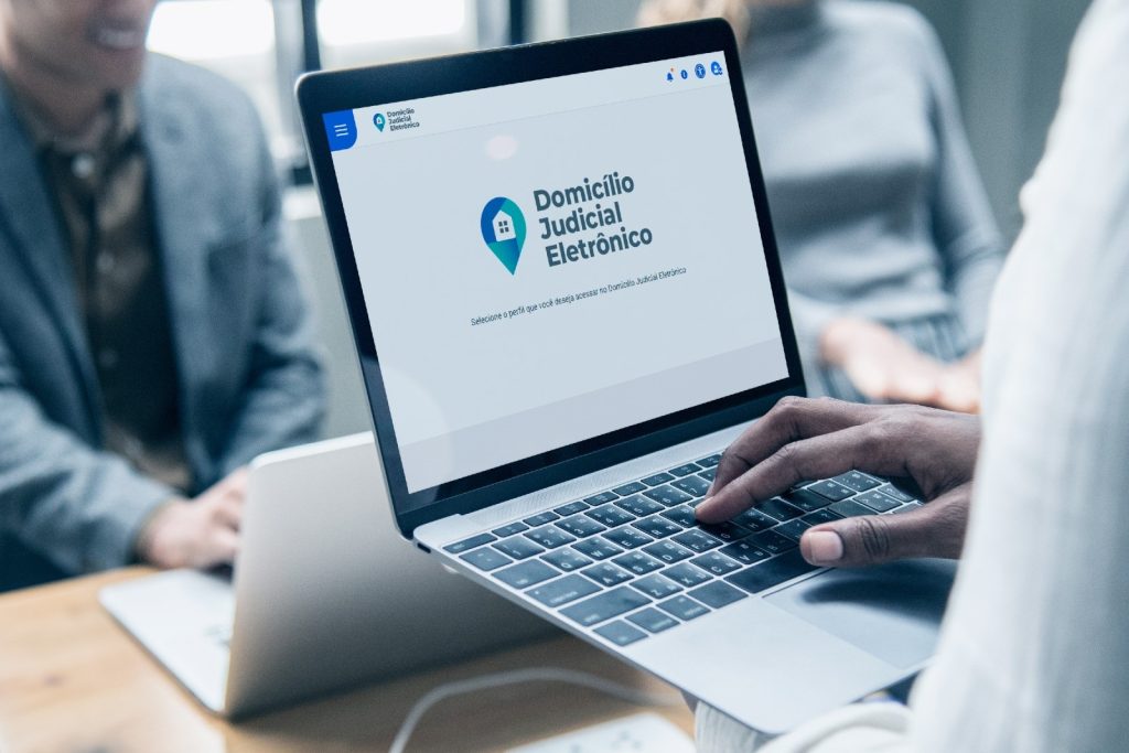 Uma pessoa mexe em um notebook em que a tela apresenta a inscrição “Domicílio Judicial Eletrônico” e a logotipo que é composta por uma casa dentro de um símbolo de localização. Outras duas pessoas aparecem ao fundo, que está desfocado. 