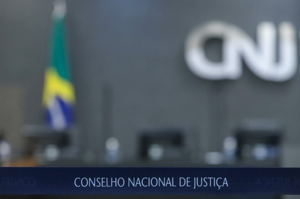 Fotografia do Plenário do CNJ, com o fundo desfocado e em destaque uma faixa azul com a inscrição “Conselho Nacional de Justiça”. É possível ver a silhueta de alguns itens ao fundo, como a bandeira do Brasil e o logotipo do CNJ na parede.  