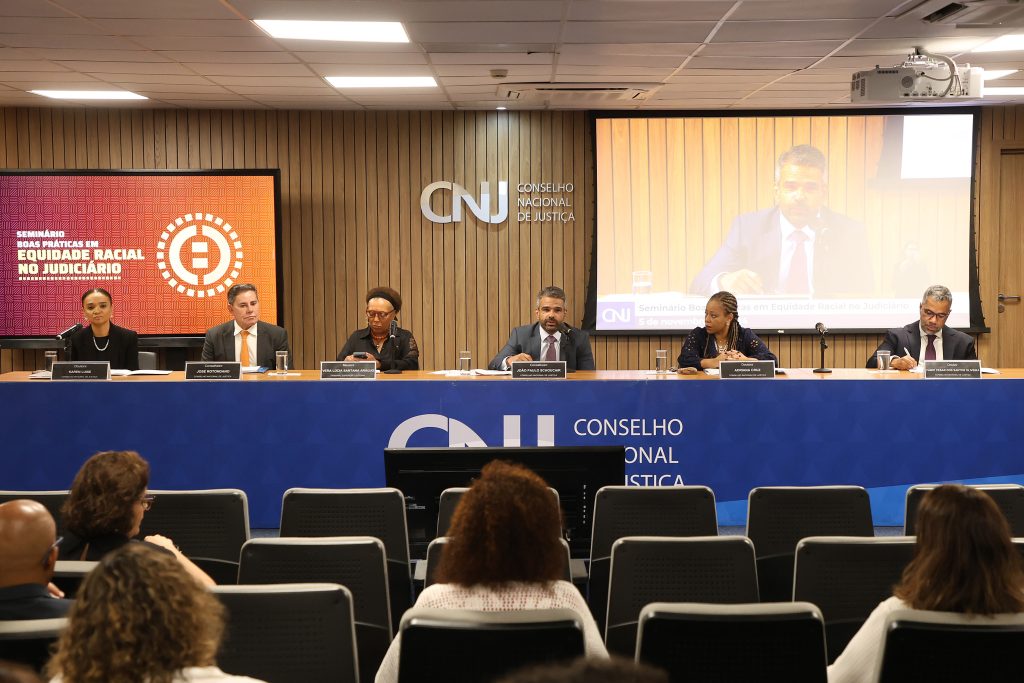 Seis pessoas sentadas atrás de uma mesa de madeira e que possui a inscrição “Conselho Nacional de Justiça” juntamente com o logotipo do CNJ estampada na frente. Três mulheres e três homens compõem a mesa. Ao fundo, atrás das pessoas, há um uma tela de projeção e uma televisão, que transmite a inscrição “Seminário Boas Práticas em Equidade Racial no Judiciário”. Outras pessoas assistem ao seminário. 