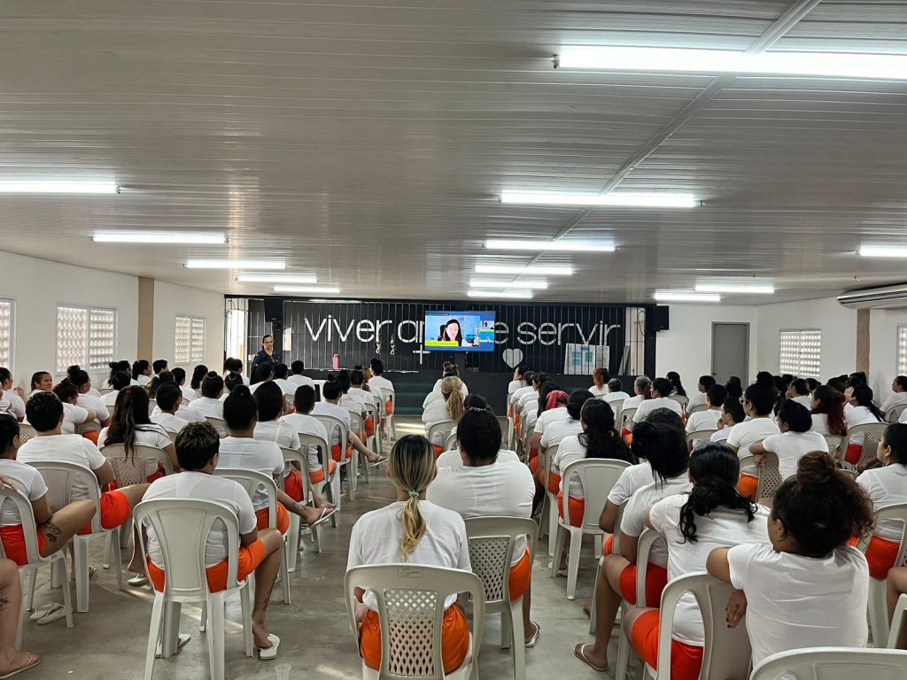 Diversas mulheres dentro de uma unidade prisional assistem à transmissão da 5.ª Jornada Literária no Cárcere. Há aproximadamente 90 pessoas sentadas, e vestem uma blusa branca e uma bermuda laranja. Elas estão sentadas em cadeiras de plástico. Em frente a elas, uma televisão transmite ao evento. Ao fundo, uma parede preta com uma frase pintada de branca*. *não é possível ver a frase por completa