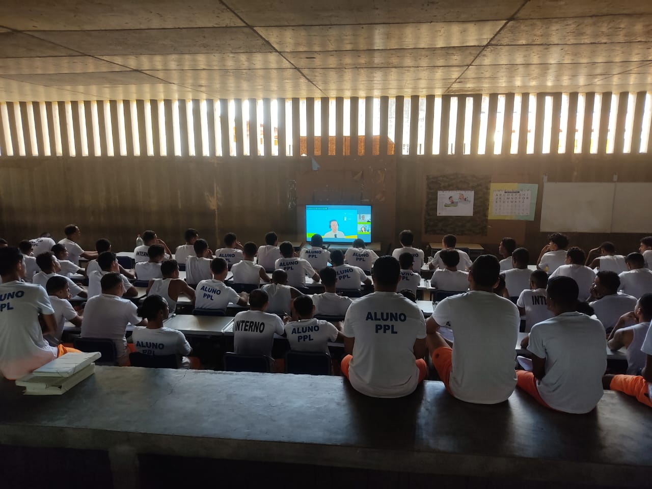 Diversos homens dentro de uma unidade prisional assistem à transmissão da 5.ª Jornada Literária no Cárcere. Há aproximadamente 50 pessoas sentadas, e vestem uma blusa branca e uma bermuda laranja. Algumas das blusas tem inscrições como “ALUNO PLL” e “INTERNO”. Em frente a eles, uma televisão transmite ao evento. 