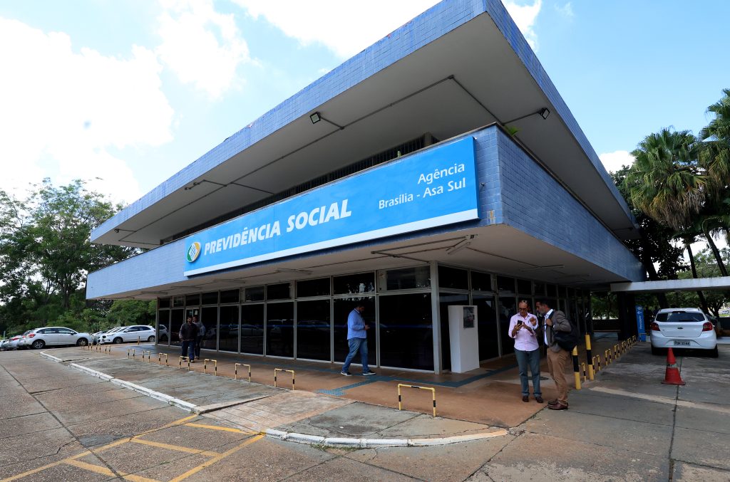 Fotografia da entrada da agência do INSS em Brasília. Prédio com fachada azul e com a inscrição “Previdência Social Agência Brasília – Asa Sul”, tem também paredes de vidro. Pessoas chegando ao local e outras estão paradas conversando. Um carro branco está estacionado próximo a porta. Ao fundo há algumas árvores e outros carros estacionados. 