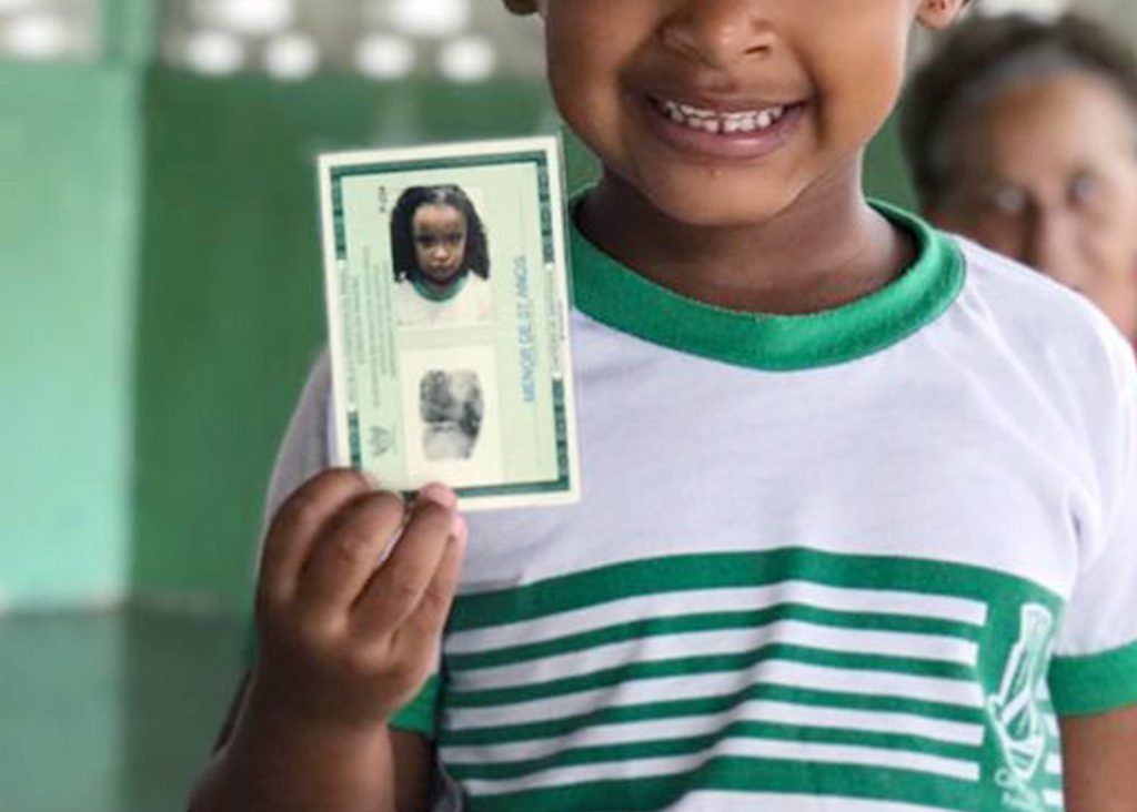 Fotografia de uma criança enquanto segura uma identidade (RG). Ela veste uma blusa branca com listras verdes. Não é possível ver seu rosto.  