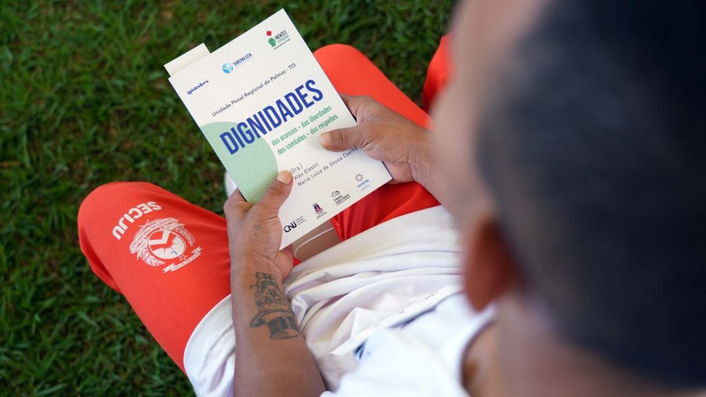 Fotografia de um homem sentado enquanto segura um livro que tem como título a inscrição “DIGNIDADES”. Ele veste uma blusa branca e bermuda laranja.