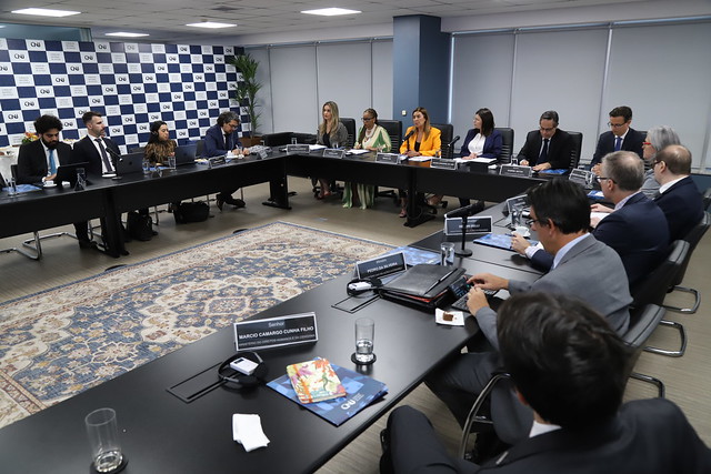 Fotografia do representante da Comissão Interamericana de Direitos Humanos (CIDH), Pedro Vaca, diversos conselheiros do CNJ, como as doutoras Leila Mascarenhas, Adriana Cruz, Renata Gil, Daiane Nogueira e o doutor Gabriel Matos e representantes de outros órgãos. Eles estão sentados atrás de uma mesa preta em formato de “U”. No lado esquerdo da foto há um painel quadriculado nas cores azul e branca, com o logotipo do CNJ. 