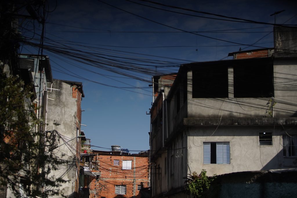 Fotografia de algumas casas de dois andares com tijolos aparentes. No lado esquerdo da foto, há algumas plantas trepadeiras nos muros. Alguns fios de energia passam entre os telhados das casas. O céu está azul.  