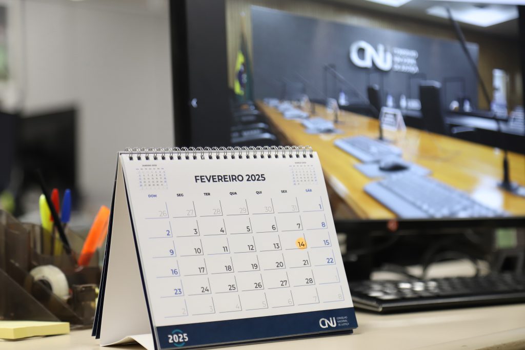 Fotografia de um calendário, na página de fevereiro, sobre uma mesa. Além dele, há um computador, que transmite uma foto do plenário do CNJ vazio, um porta canetas, com marca textos, canetas, uma fita pequena e um bloco de post it.  