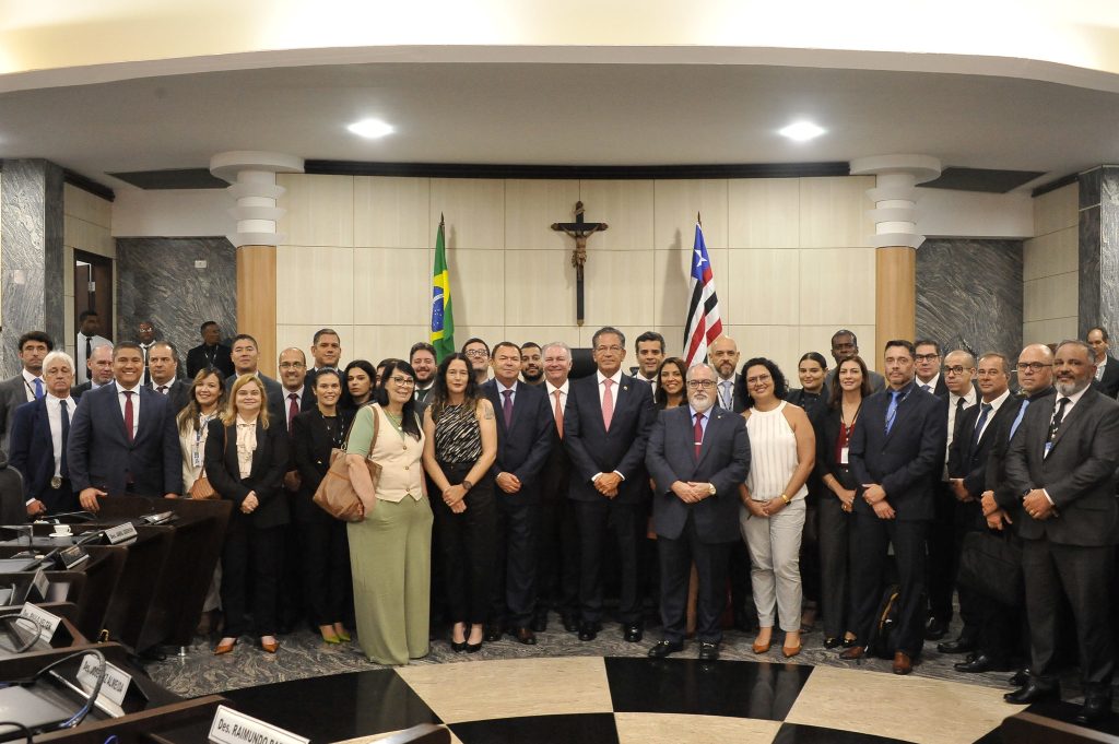 Fotografia de diversas pessoas, entre eles o ministro Mauro Campbell, em frente a dois mastros com bandeiras do Brasil e do Maranhão e de um crucifixo que está na parede ao fundo. Eles estão no Tribunal de Justiça do Maranhão (TJMA). 