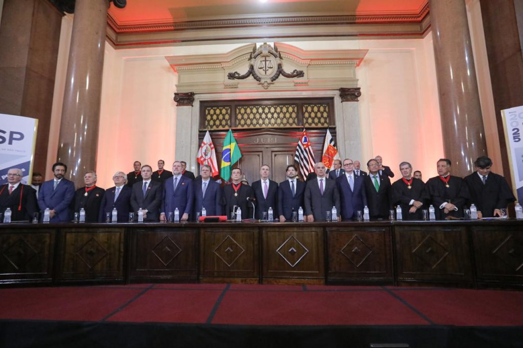 Fotografia do presidente do Conselho Nacional de Justiça (CNJ) e do Supremo Tribunal Federal (STF), ministro Luís Roberto Barroso e de mais outros vinte e três homens no Tribunal de Justiça de São Paulo (TJSP). Eles estão em pé atrás de uma mesa de madeira e vestem ternos e togas. Ao fundo, atrás deles, há uma porta de madeira e quatro mastros com bandeiras, como a do Brasil e a do Estado de São Paulo.  